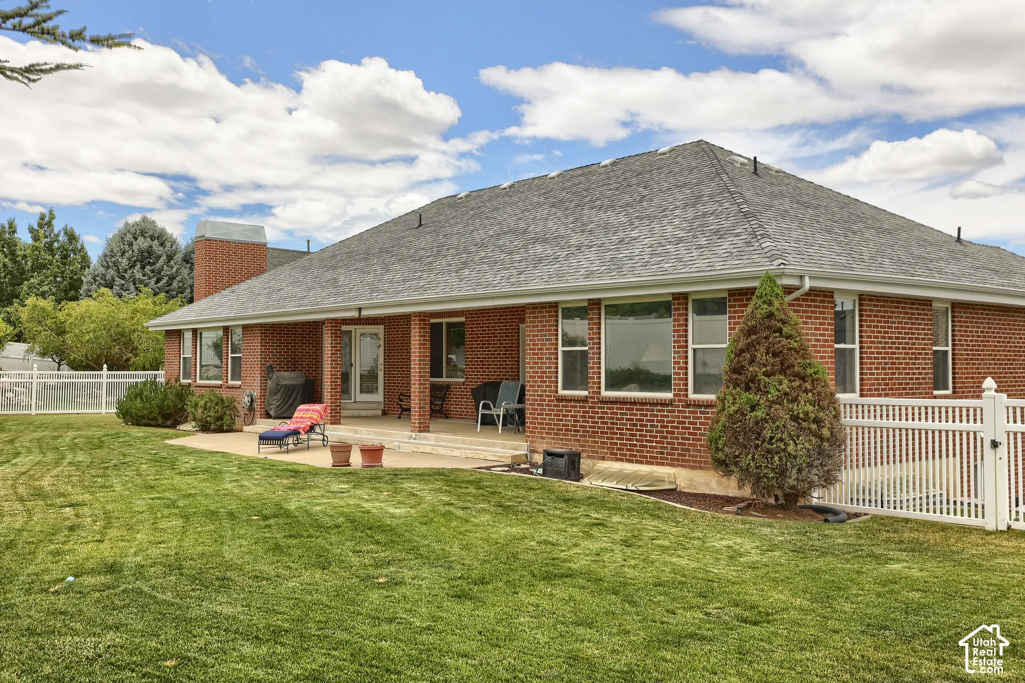 Expansive green lawn bordering a brick exterior with a covered patio area, featuring multiple windows and a white picket fence