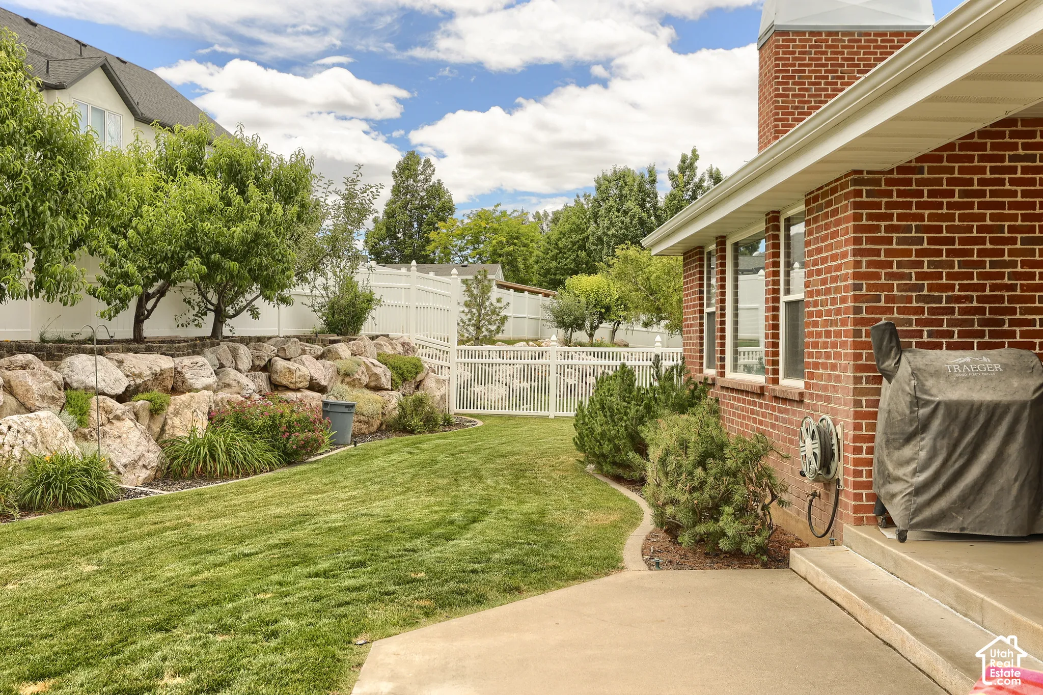 Expansive backyard featuring a manicured lawn, a rock retaining wall, mature trees, and a white privacy fence
