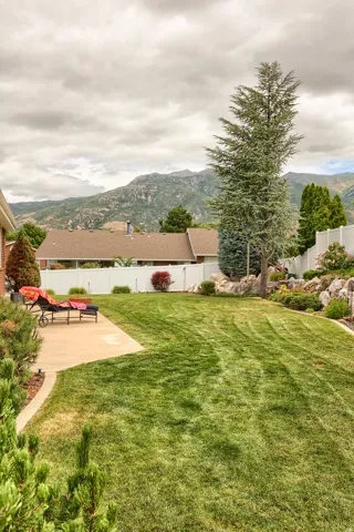 Expansive backyard featuring a lush lawn, concrete patio, mature evergreen trees, and a white privacy fence
