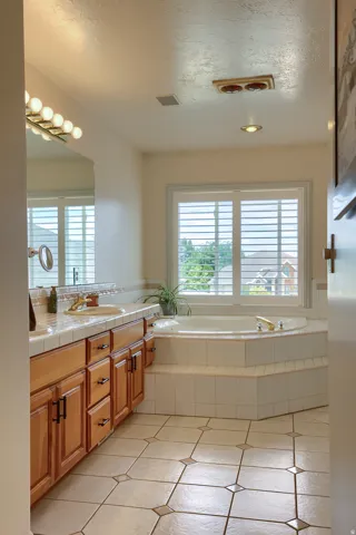 Corner soaking tub with a tiled surround, plantation shutters, and an adjacent vanity with wood cabinetry and a tile countertop