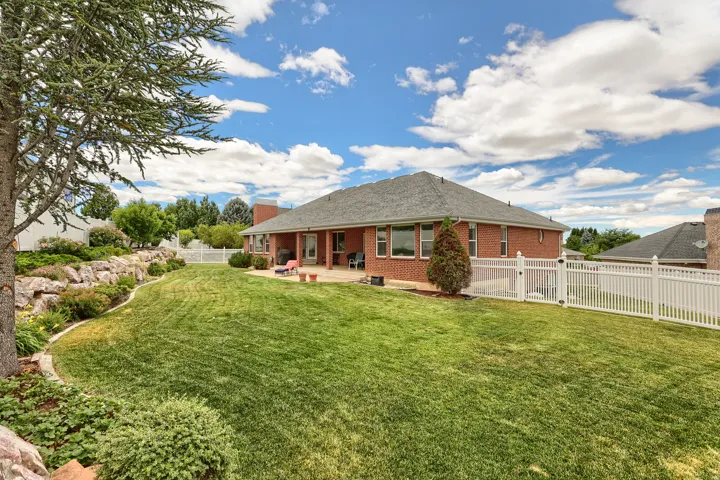 Expansive backyard featuring a manicured lawn, a white picket fence, a rock retaining wall with mature landscaping, a paved patio, and a brick exterior with multiple windows