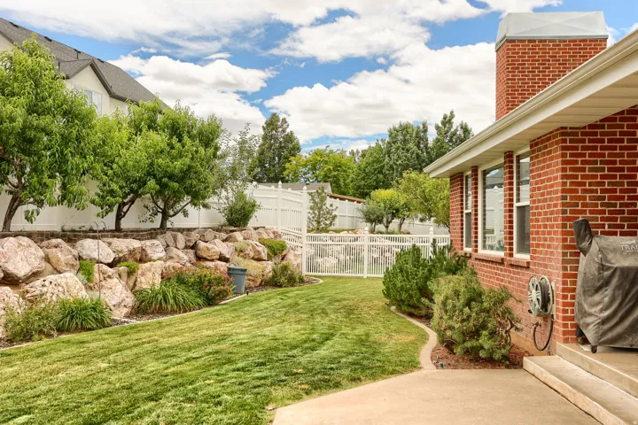 Expansive backyard featuring a lush lawn, a concrete patio, a red brick exterior, and a rock retaining wall with mature trees and shrubbery