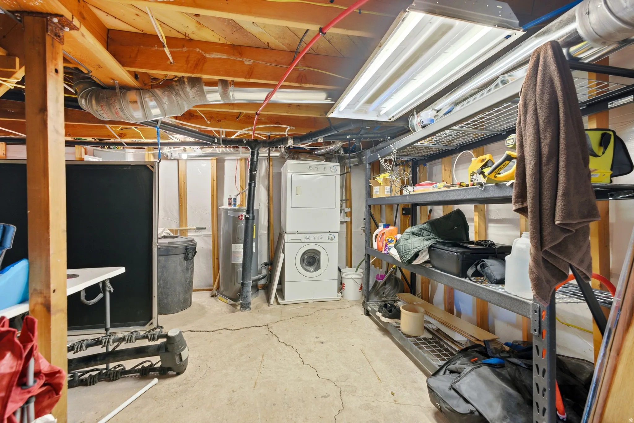 Utility space featuring exposed ceiling joists, a stacked washer and dryer, a water heater, and metal shelving units