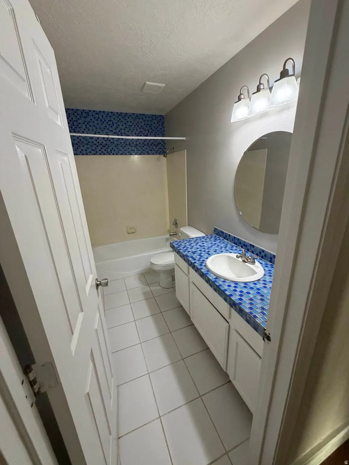 Bathroom featuring a white vanity with a blue mosaic tile countertop, an integrated white sink, and a circular wall mirror