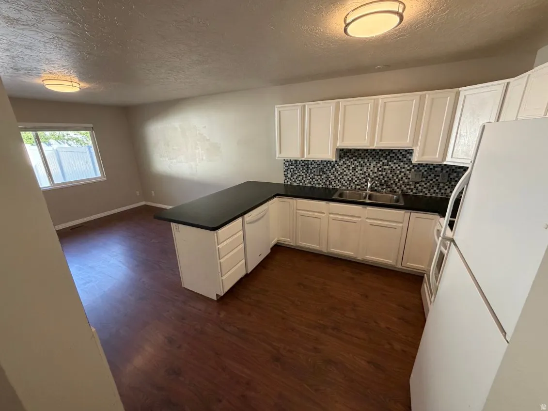 Kitchen area featuring dark wood-finish flooring, white cabinetry, dark countertops, a mosaic tile backsplash, and a double basin sink