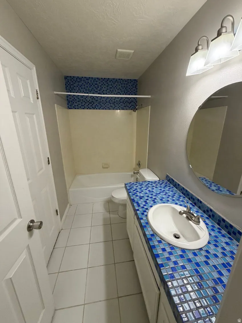 Bathroom featuring a white vanity with a blue mosaic tile countertop, an oval-shaped mirror, a white drop-in sink with a brushed nickel faucet, a white toilet, and a white bathtub with blue mosaic tile surround