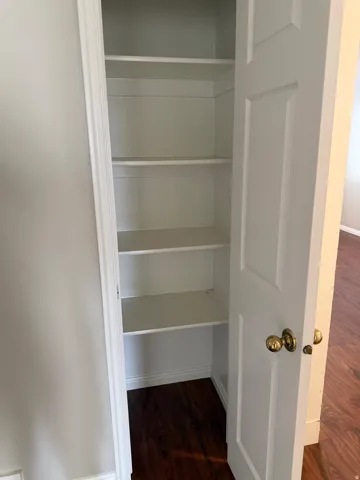 Built-in storage closet featuring multiple white shelving units, a white paneled door with brass-tone hardware, and dark wood-finish flooring