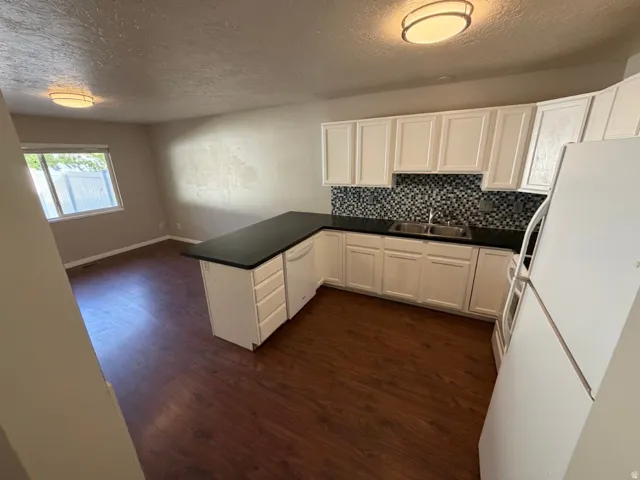 Kitchen area featuring dark wood-finish flooring, white cabinetry, dark countertops, a mosaic tile backsplash, and a double basin sink