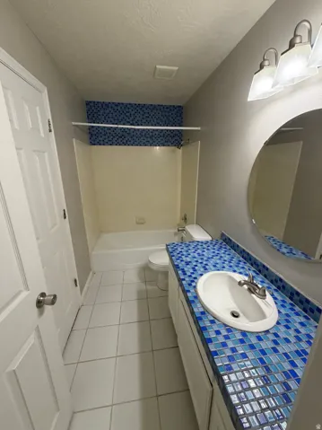 Bathroom featuring a white vanity with a blue mosaic tile countertop, an oval-shaped mirror, a white drop-in sink with a brushed nickel faucet, a white toilet, and a white bathtub with blue mosaic tile surround