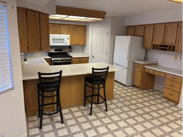Wood-finish cabinetry, built-in desk area, recessed lighting, white appliances, and a breakfast bar