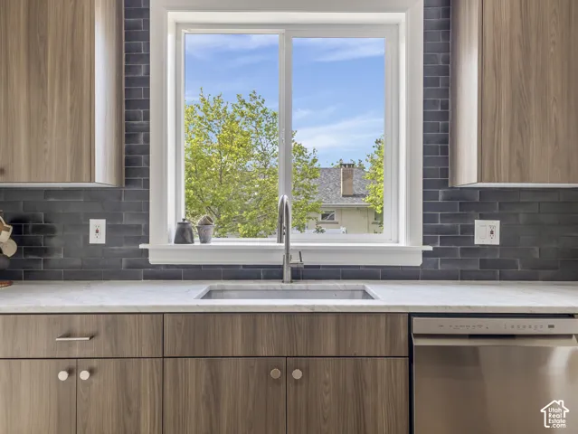 Home 1/2 -- 1046 S Lincoln Street -- Kitchen featuring a stainless steel sink with a gooseneck faucet, light-colored countertop, dark subway tile backsplash, wood-finish cabinetry with various hardware, and a built-in stainless steel dishwasher