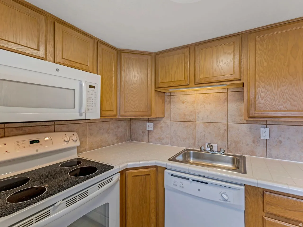 Kitchen featuring light wood cabinetry, white appliances, tile countertops, and a tiled backsplash