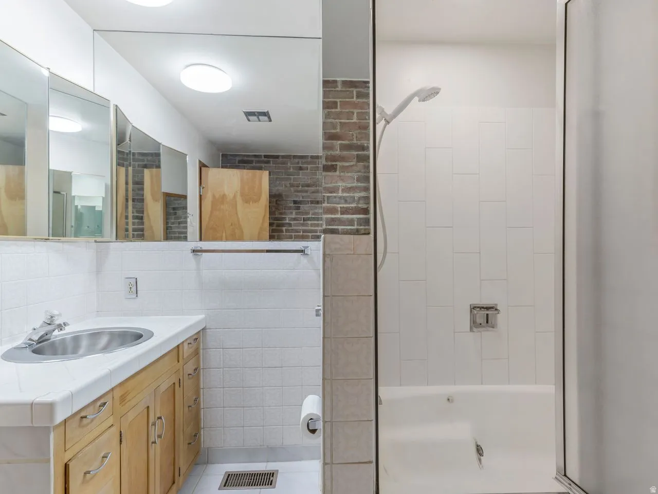 Bathroom featuring a light wood-finish vanity with an integrated sink, a large wall-mounted mirror, and a full bathtub with a shower head and white tiled surround