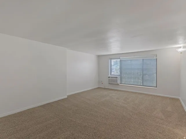 Spacious room featuring neutral carpeting, white walls, a large window with horizontal blinds, and a built-in air conditioning unit