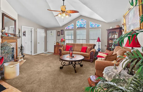 Vaulted ceiling with a ceiling fan, carpeted flooring, a fireplace with a wood mantle, and a series of windows featuring decorative glass accents