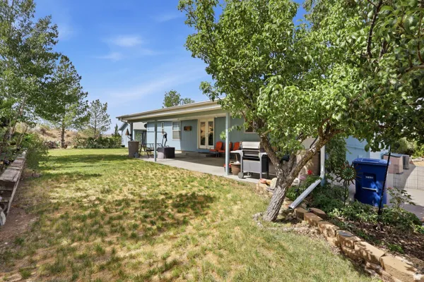 Expansive backyard featuring a covered patio, a manicured lawn area, mature trees, and a retaining wall