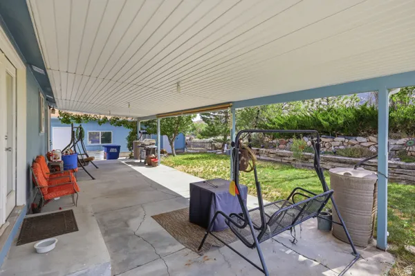 Expansive covered patio with concrete flooring, supported by columns, offering direct access to the landscaped yard with mature shrubbery and a stone retaining wall