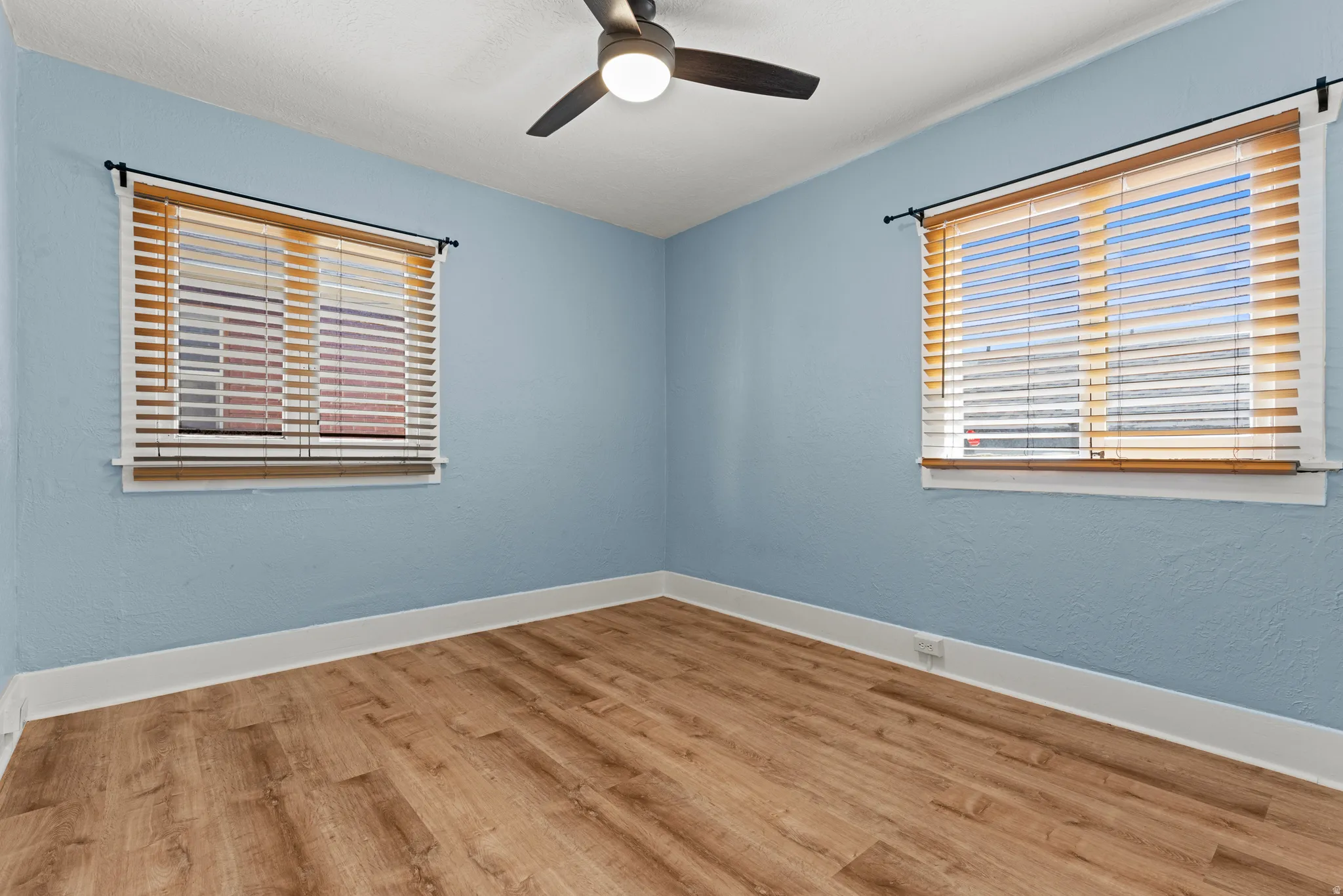 Interior room featuring wood-finish flooring, two windows with blinds, a ceiling fan with integrated lighting, and white baseboards