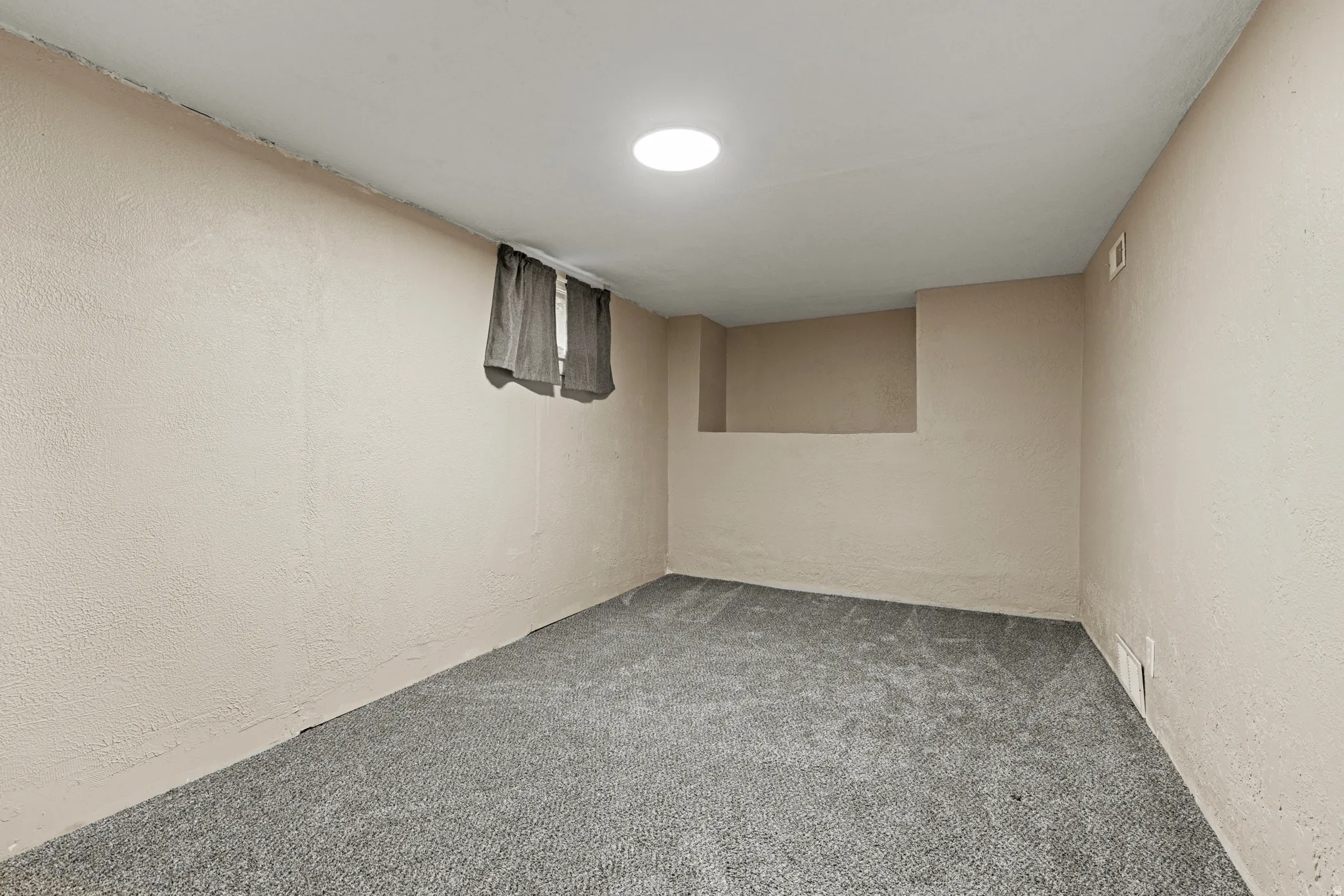 Carpeted room with a recessed circular light fixture, a small window with drapes, and a wall protrusion