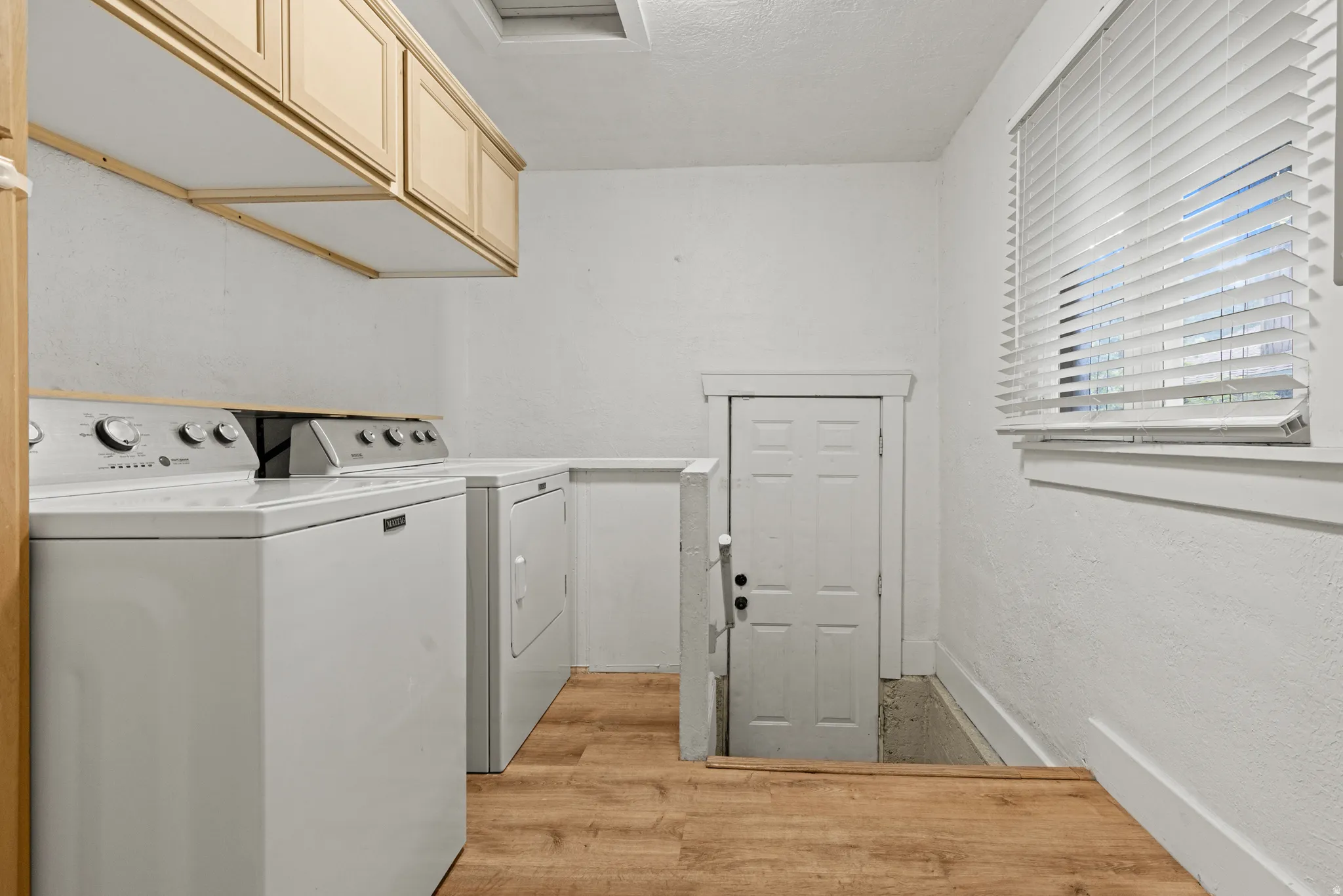Dedicated laundry area featuring wood-finish flooring, overhead cabinetry, and a window with horizontal blinds