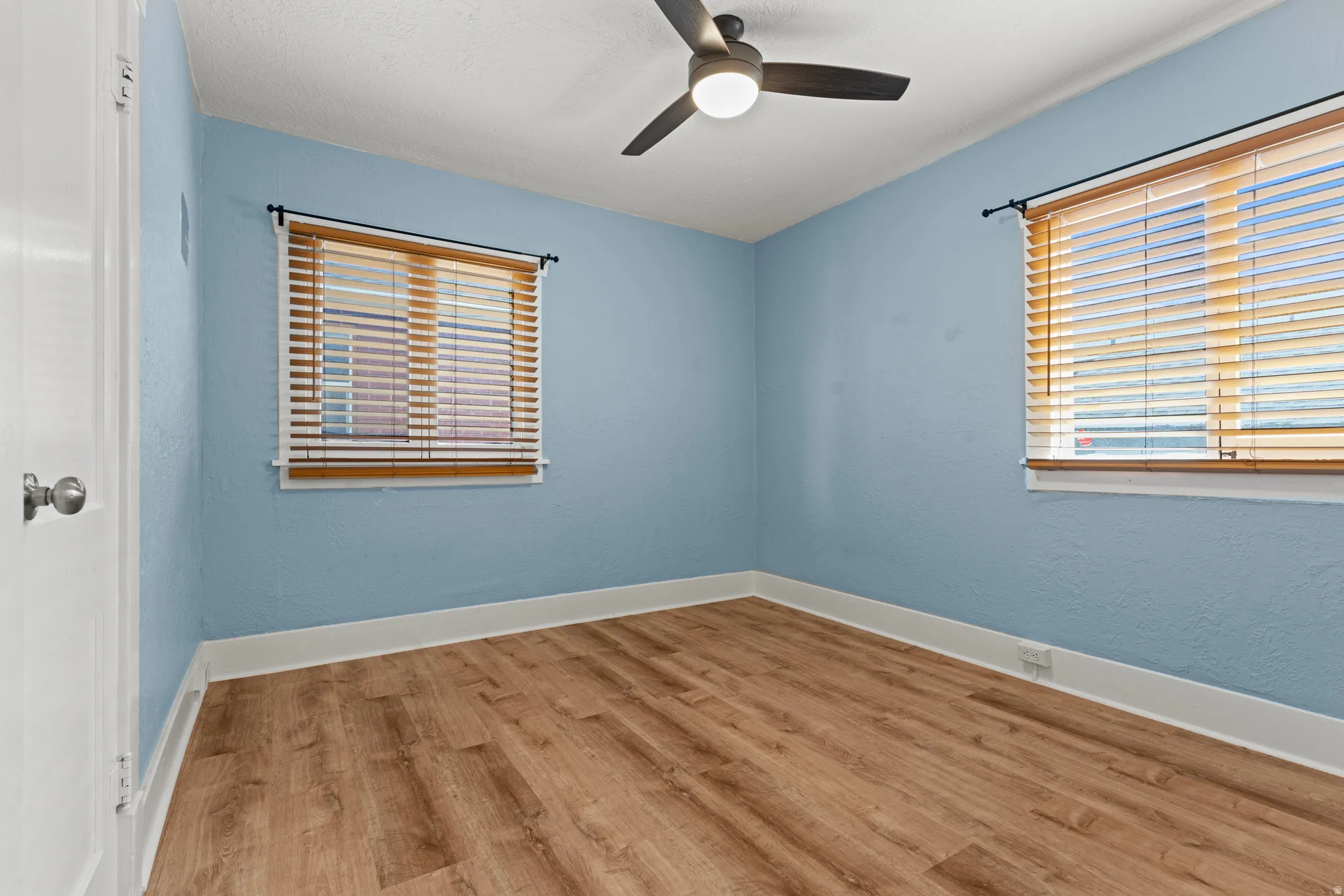 Room featuring wood-finish flooring, two windows with wooden blinds, a ceiling fan with integrated lighting, and crisp white baseboards