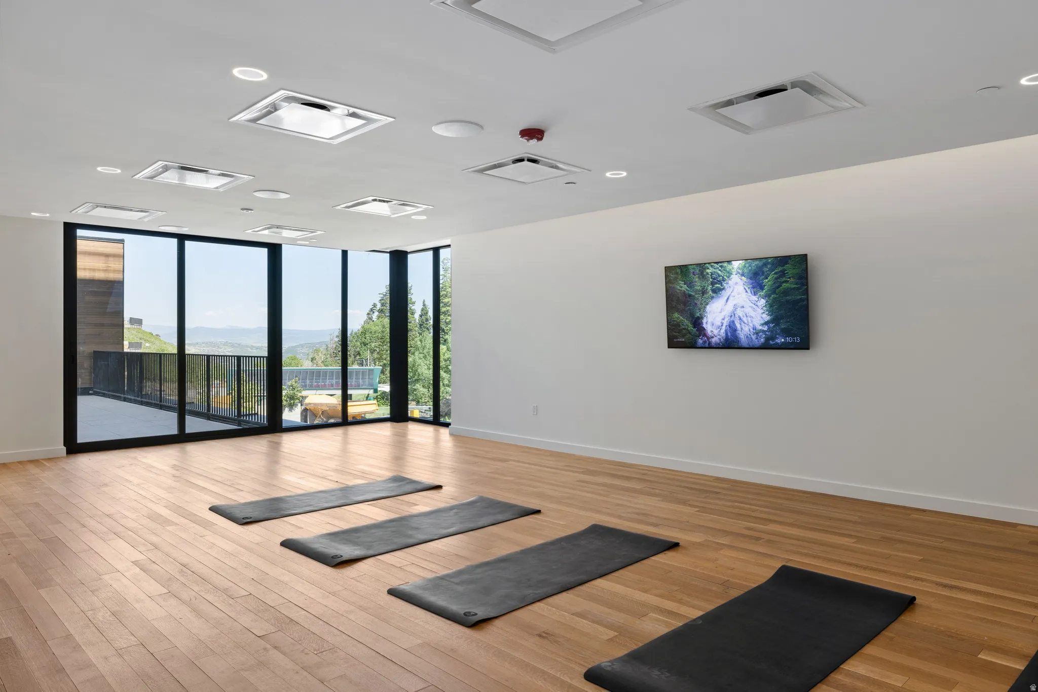 Spacious room featuring wood-finish flooring, floor-to-ceiling windows, multiple recessed ceiling lights, and a wall-mounted display