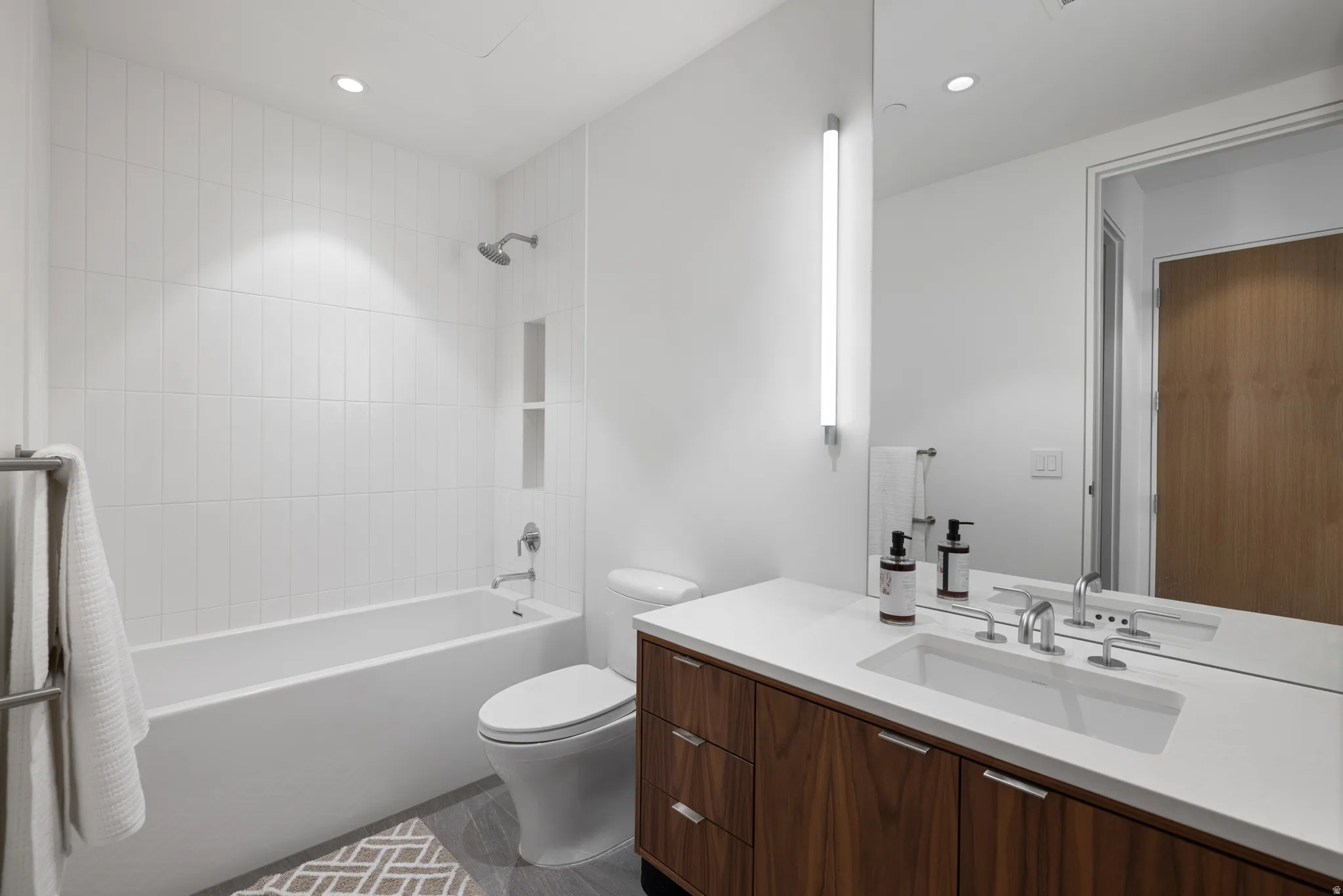 Contemporary bathroom featuring a white subway tile tub surround, built-in shower niche, wood-finish vanity with an integrated sink, and an illuminated vertical mirror