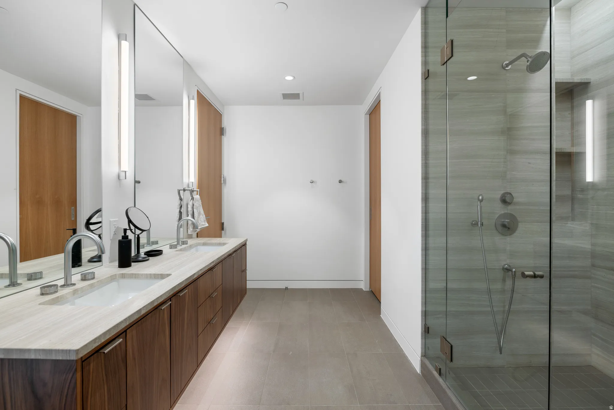 Spacious bathroom featuring a dual vanity with a light-toned countertop, wood-finish cabinetry, and a large frameless mirror