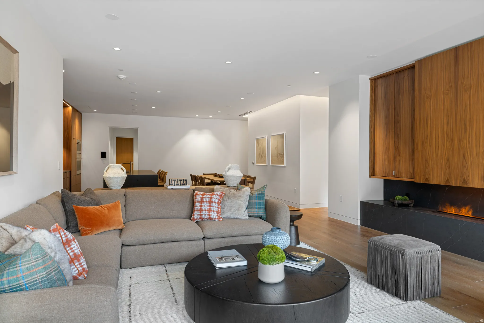 Open-concept living area featuring a modern fireplace with a black stone hearth, recessed ceiling lighting, wood-finish flooring, and a dining area visible in the background