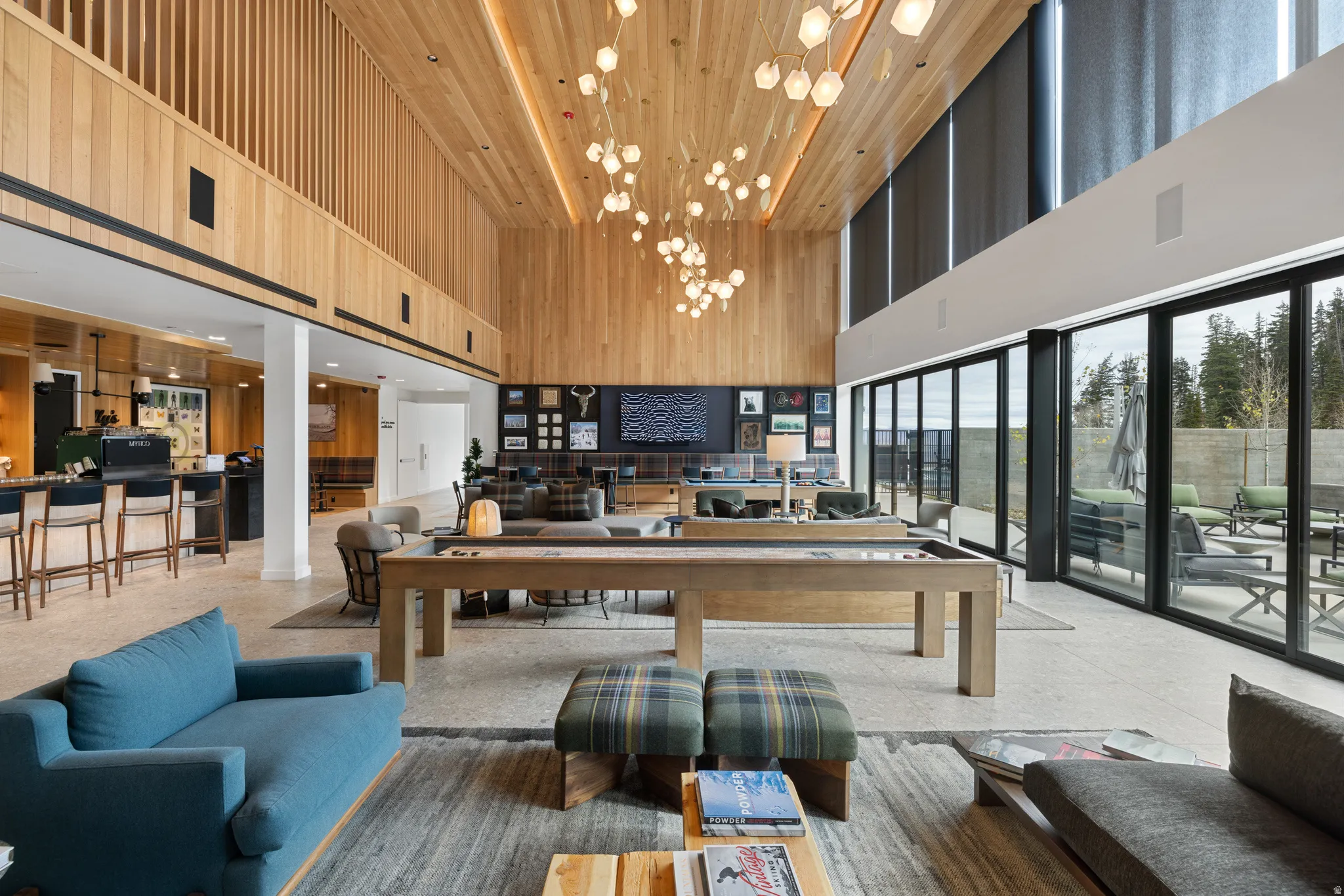 Spacious common area featuring a high vaulted ceiling with wood paneling, extensive window walls, a built-in bar with bar seating, and a shuffleboard table