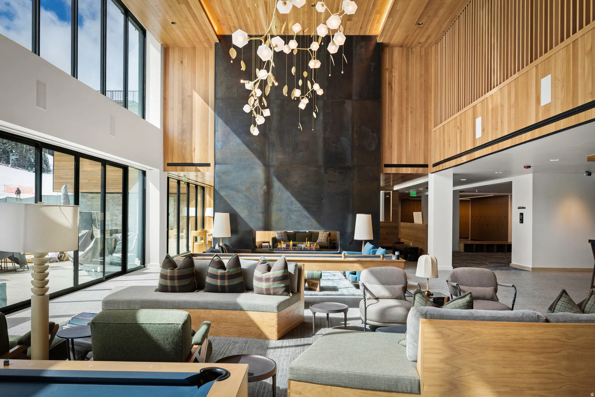 Expansive lounge featuring a dramatic two-story fireplace with dark paneling, wood-paneled walls and ceiling, a contemporary chandelier, a wall of sliding glass doors, and a built-in bench seating arrangement