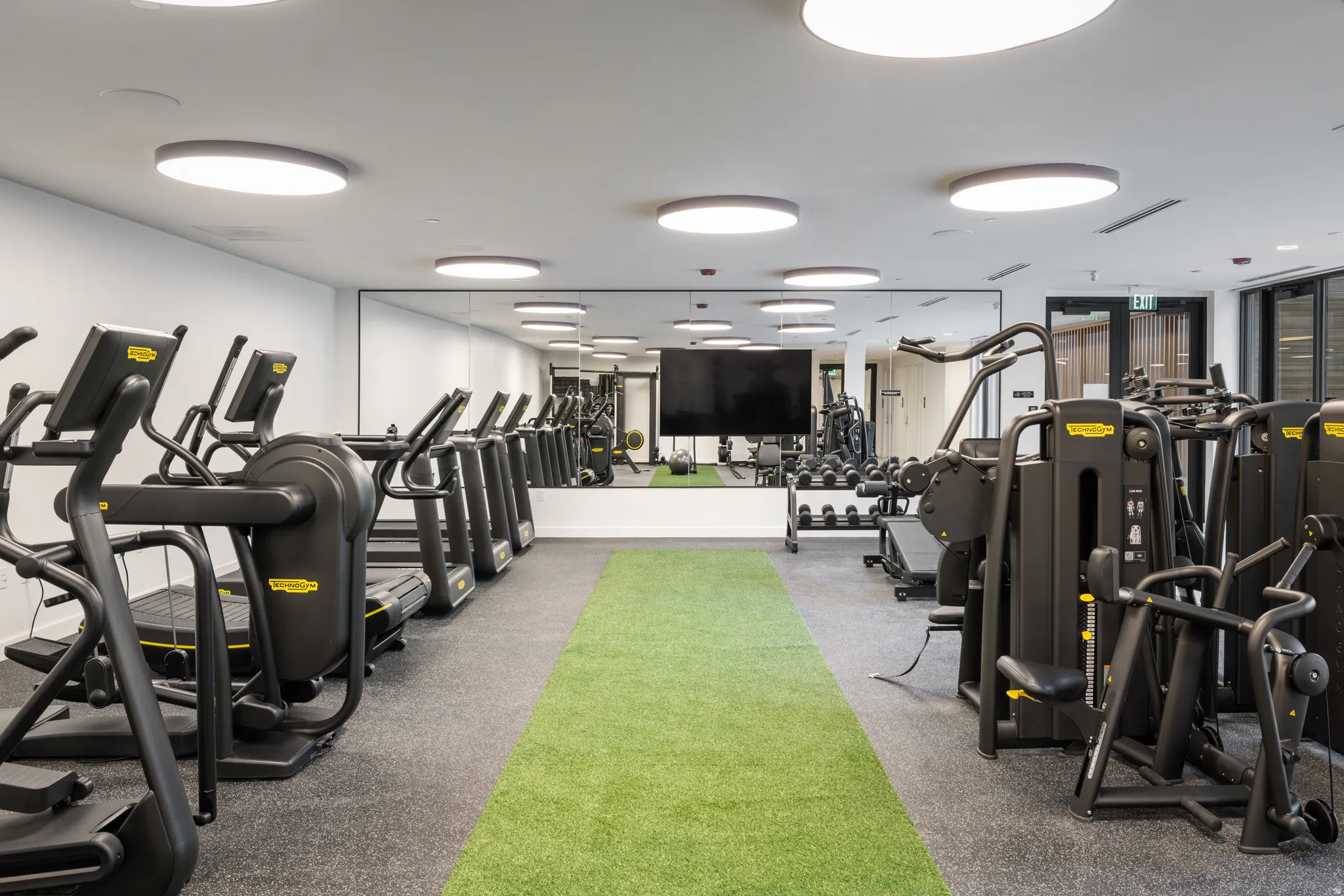 Fitness center featuring a row of cardio equipment, strength training machines, extensive mirrored wall, recessed circular lighting, and a green turf track