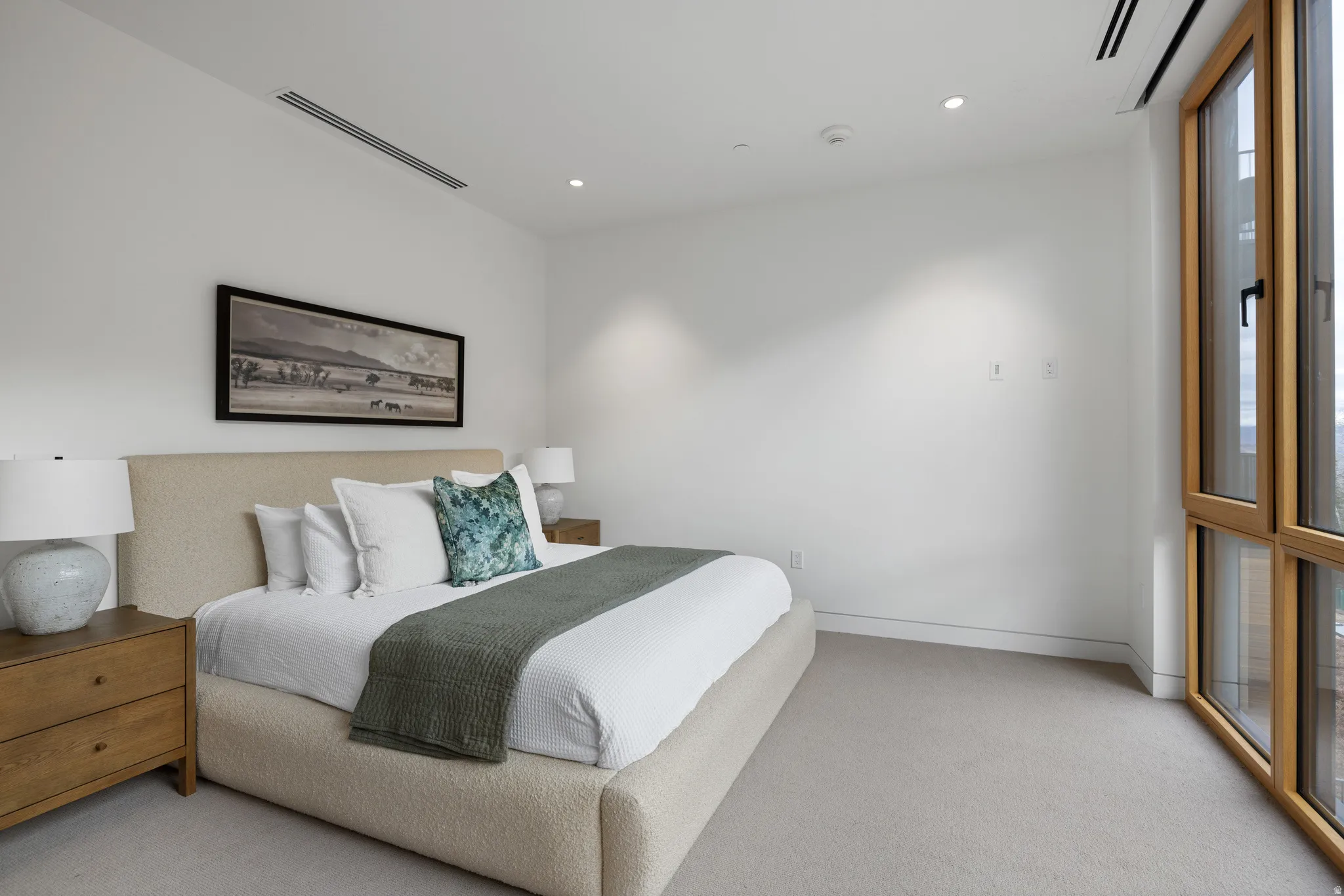 Bright bedroom featuring a large window with natural wood-finish trim, recessed lighting, linear air vents, and light carpet flooring