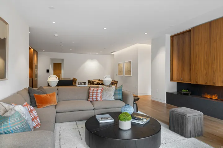 Open-concept living area featuring a modern fireplace with a black stone hearth, recessed ceiling lighting, wood-finish flooring, and a dining area visible in the background