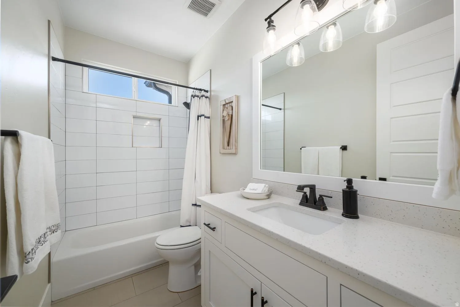 Bathroom featuring a white vanity with an integrated sink and light-colored countertop, matte black fixtures, a framed mirror with overhead lighting, a white subway-tiled tub/shower combination with a built-in niche, and neutral-toned floor tiling