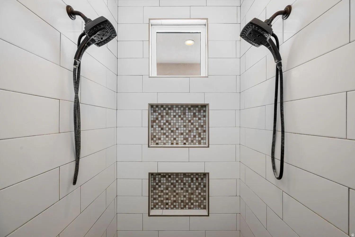 Shower with white subway tile, dual dark bronze showerheads, two recessed mosaic tile niches, and an upper transom window