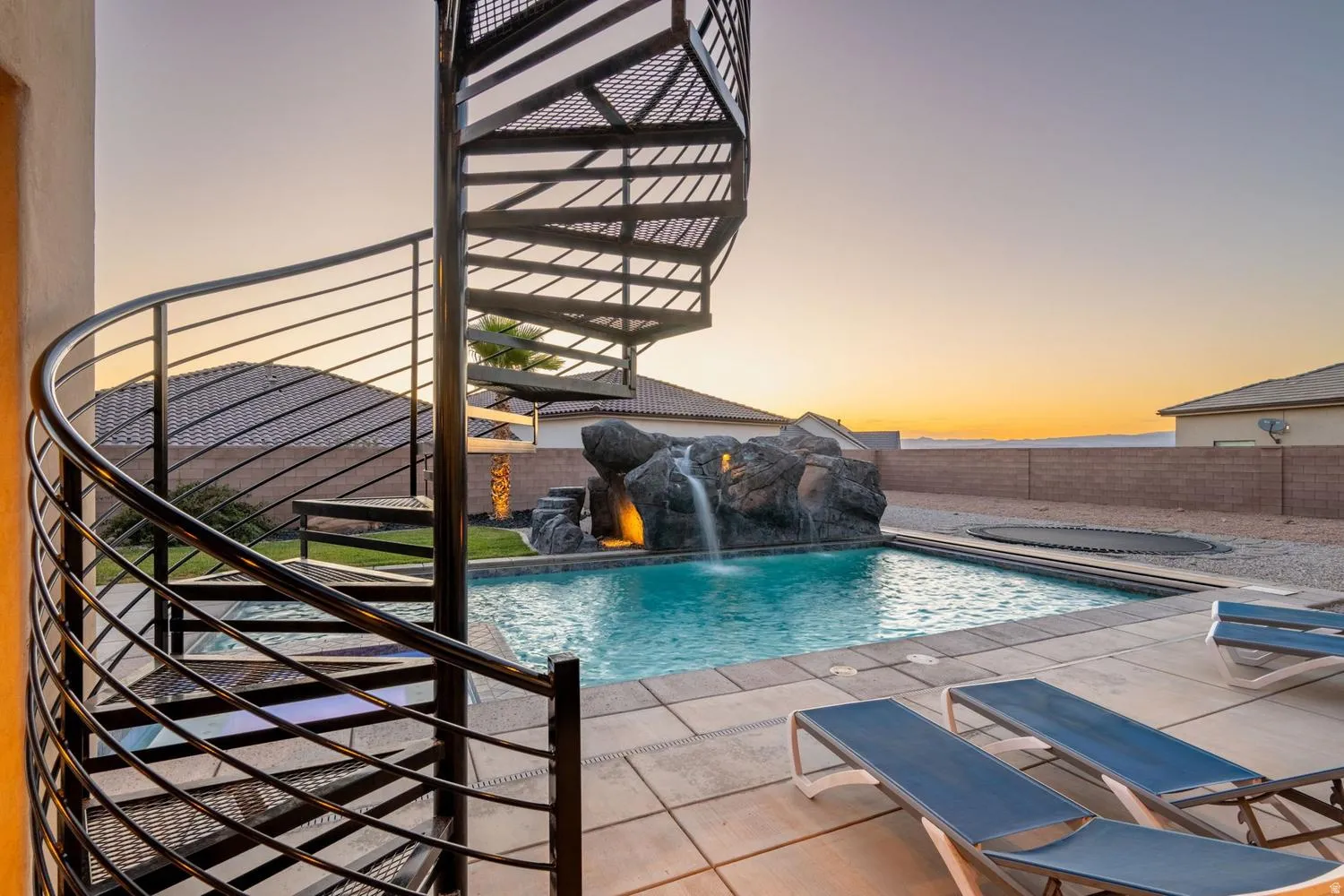 Spiral metal staircase overlooking a swimming pool with a rock waterfall feature, integrated lighting, and a surrounding paver patio
