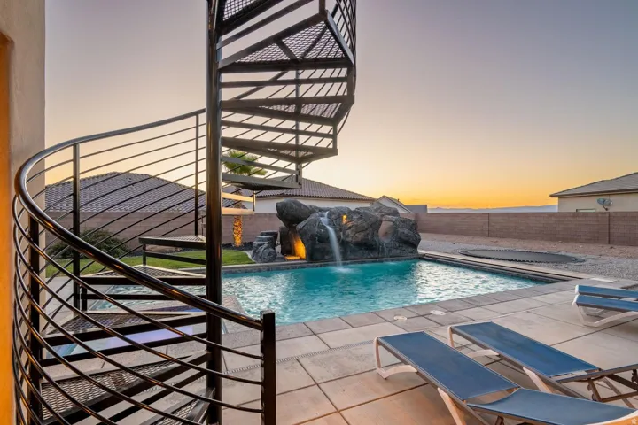 Spiral metal staircase overlooking a swimming pool with a rock waterfall feature, integrated lighting, and a surrounding paver patio