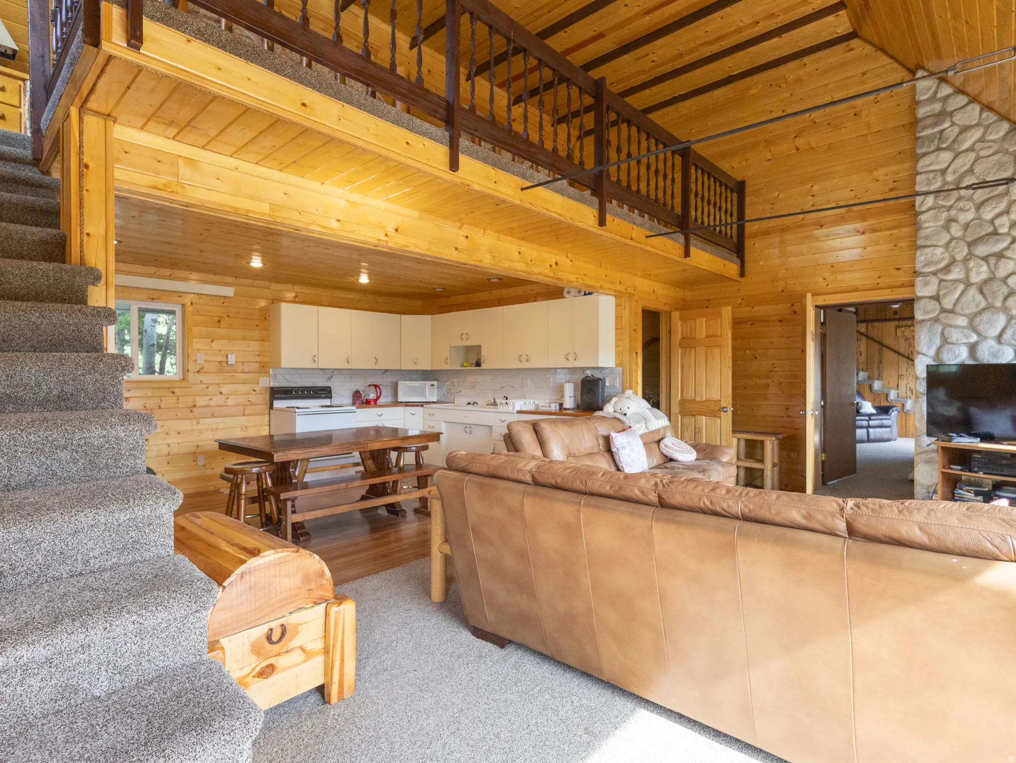 Expansive open-concept living area featuring natural wood paneling throughout, a carpeted staircase leading to a loft space with wood railing, a stone-clad column, and a kitchen area with white cabinetry and a white subway tile backsplash