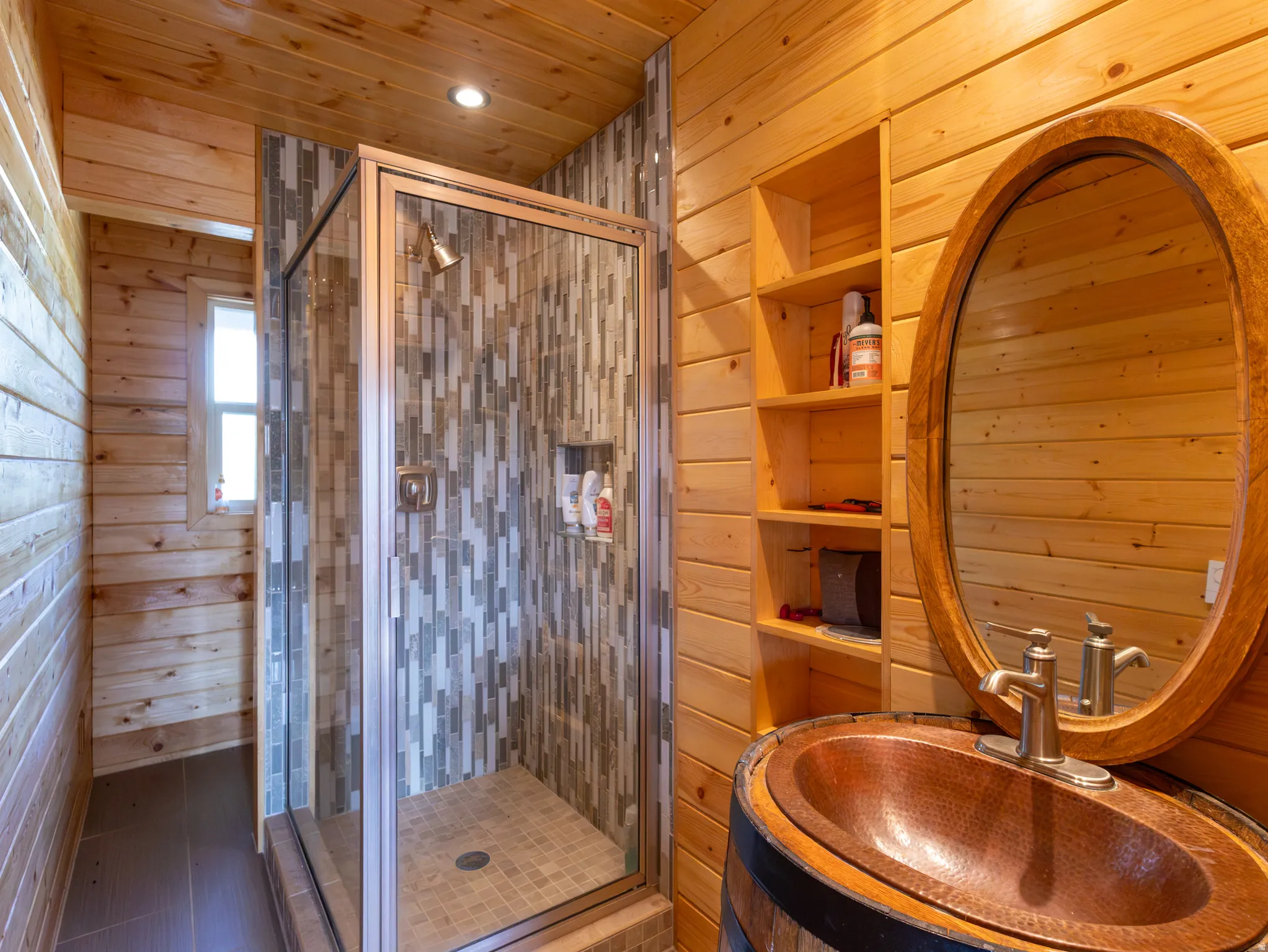 Bathroom featuring wood-finish walls and ceiling, a glass-enclosed shower with mosaic tile surround, a recessed shower niche, an oval-framed mirror, and a copper vessel sink set within a barrel-style vanity