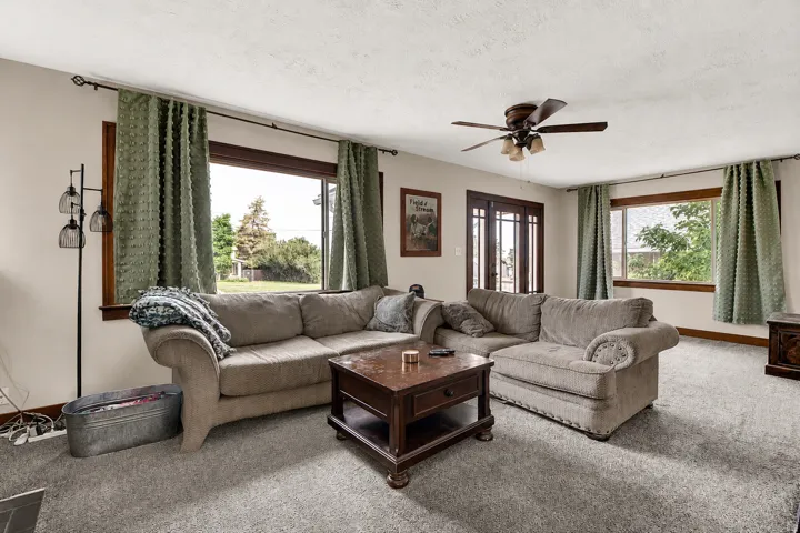Spacious living area featuring neutral carpeting, a ceiling fan with integrated lighting, natural wood trim around windows, and double French doors with transom windows