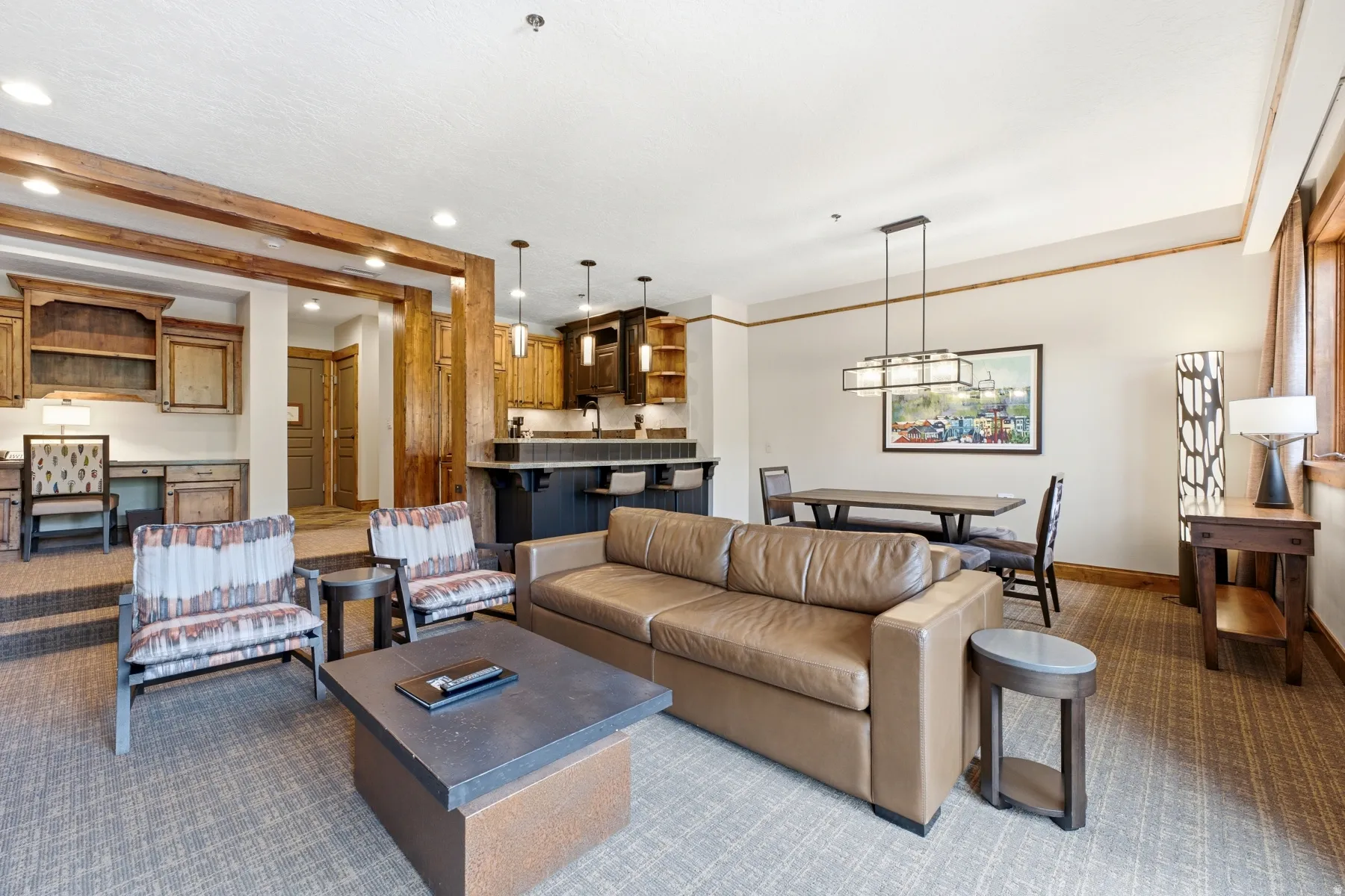 Open-concept living space featuring wood-finish ceiling beams, an integrated kitchen bar with pendant lighting, a dedicated dining area with a linear chandelier, a built-in desk with cabinetry, and textured carpeting