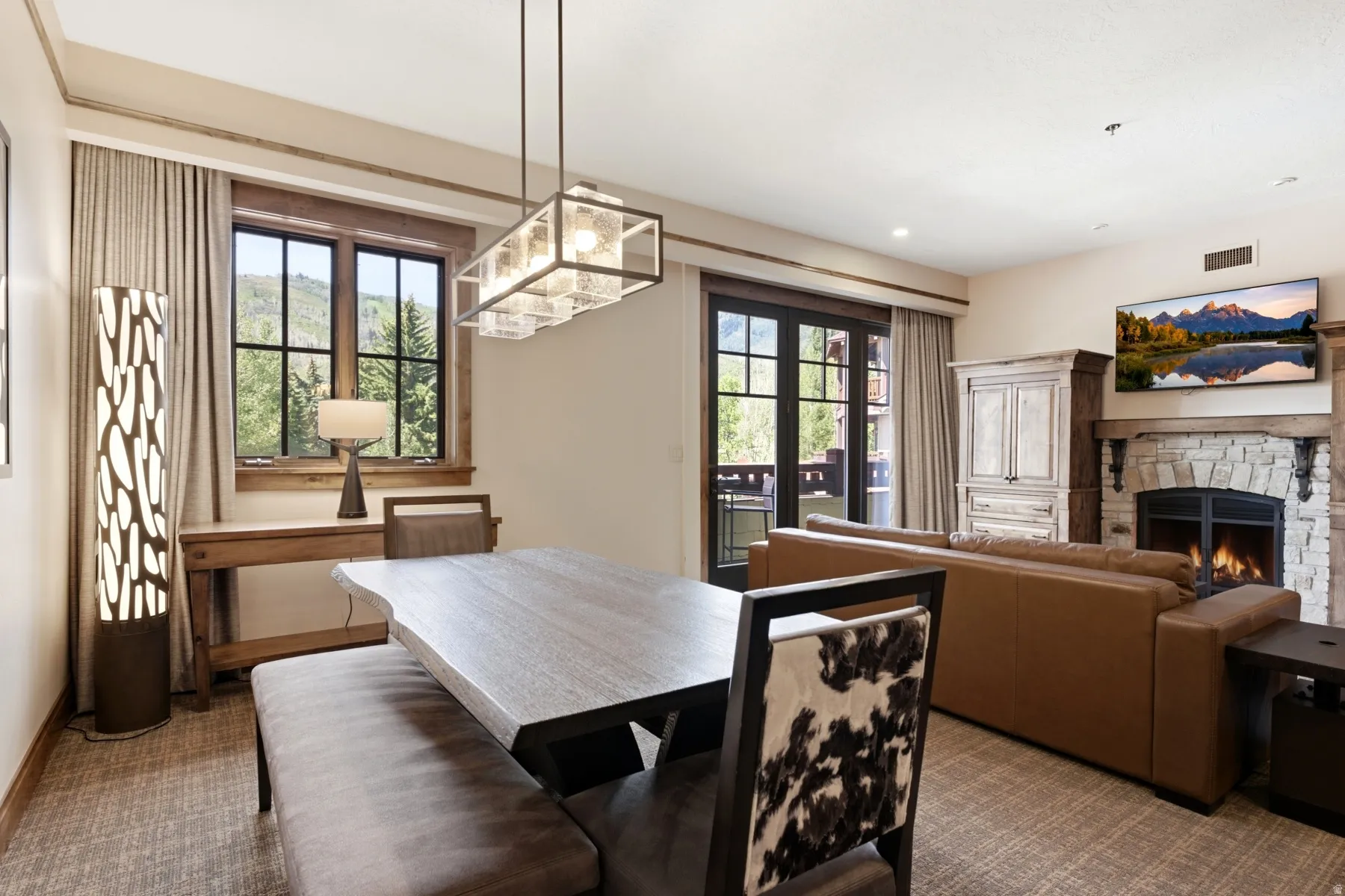 Open-concept living area featuring a stone-faced fireplace with a wood mantel, a built-in wood-finish armoire, a balcony with double glass doors, a dining area with a linear pendant light, and a window with wood trim