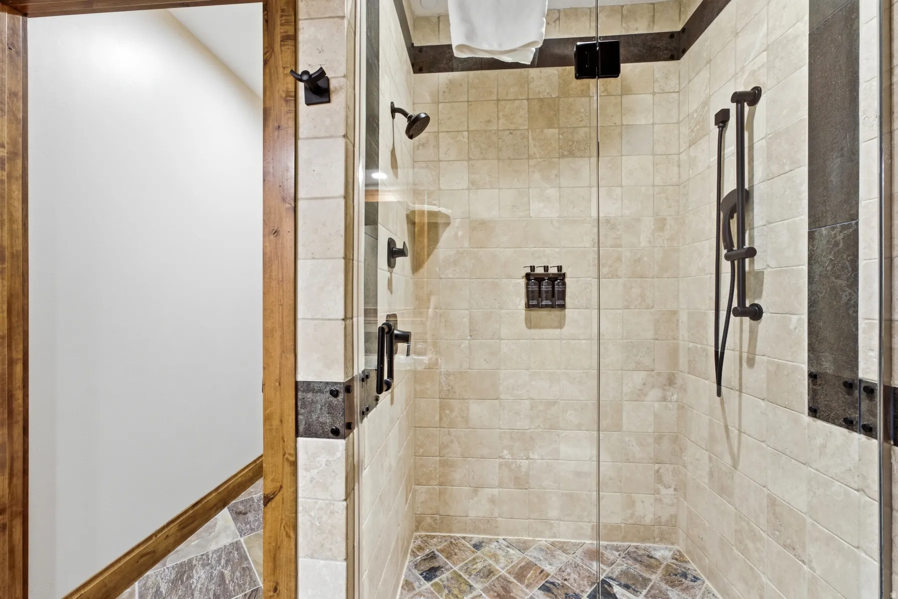 Glass-enclosed shower featuring natural stone-look wall tiles, a multi-tone stone tile shower pan, and oil-rubbed bronze fixtures including a rain shower head and a handheld sprayer