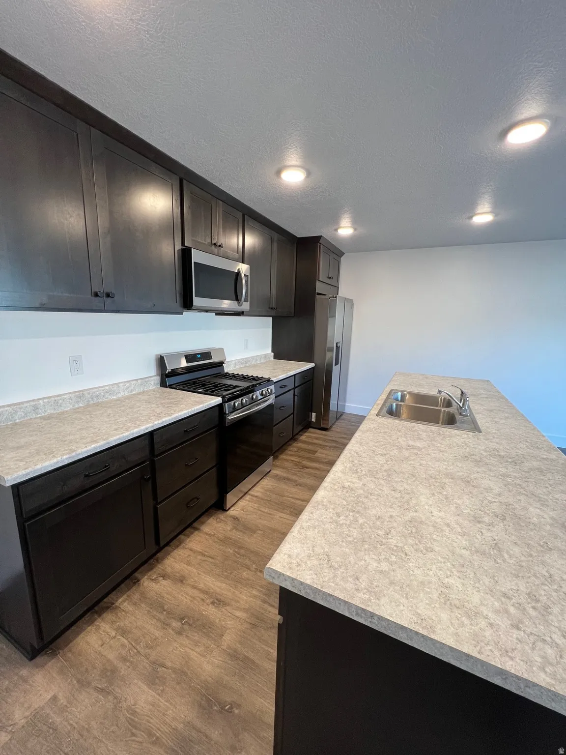 Kitchen featuring dark wood-finish cabinetry, stainless steel appliances, wood-finish flooring, recessed lighting, and light-toned countertops with an integrated double sink