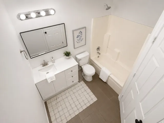 Bright bathroom featuring a single-sink vanity with white cabinetry and a rectangular mirror, a toilet, a full bathtub with an integrated shower, and dark gray tile flooring