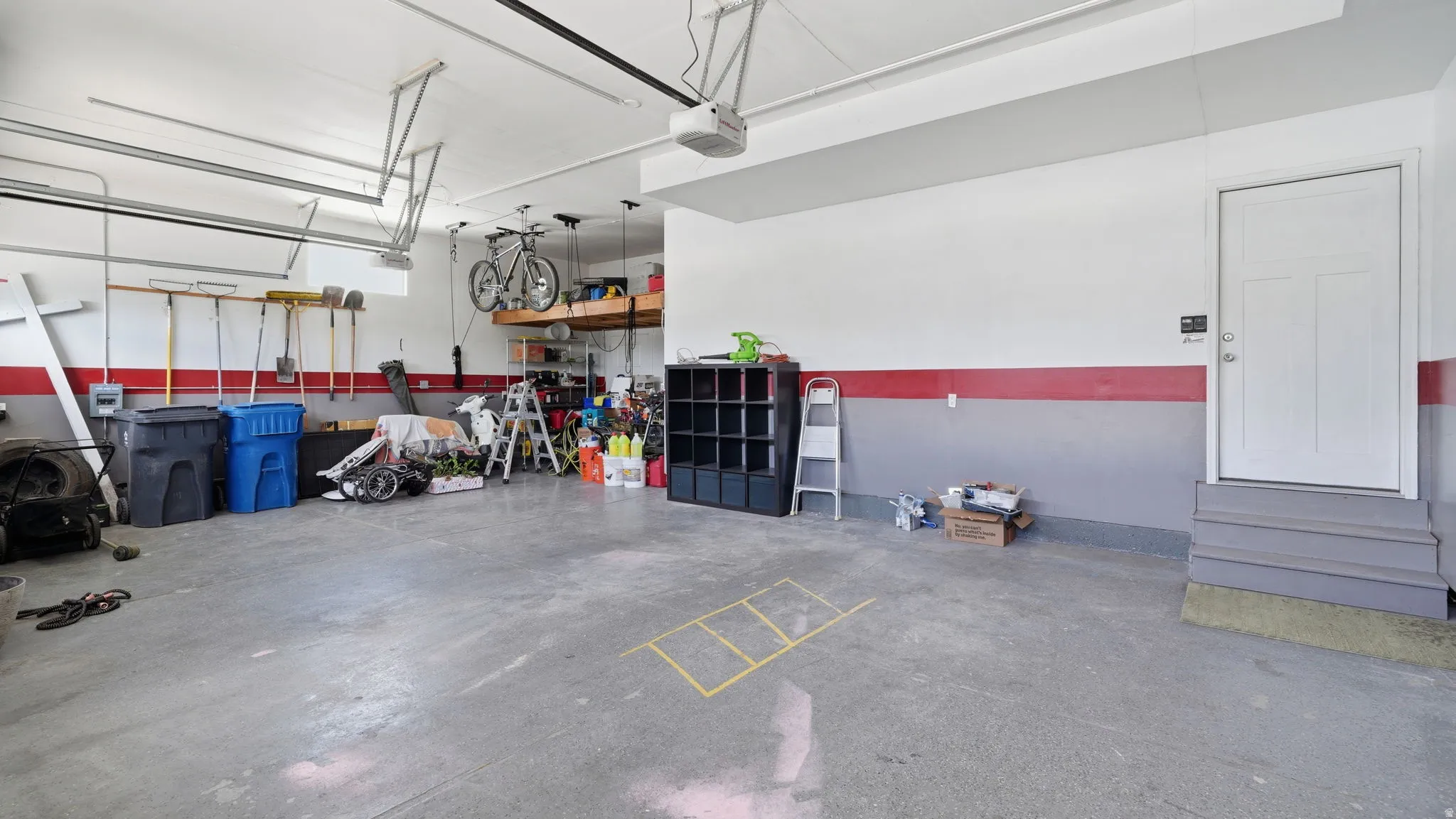 Spacious garage featuring a finished concrete floor, painted walls with a red accent stripe, an interior access door with steps, and overhead storage shelving