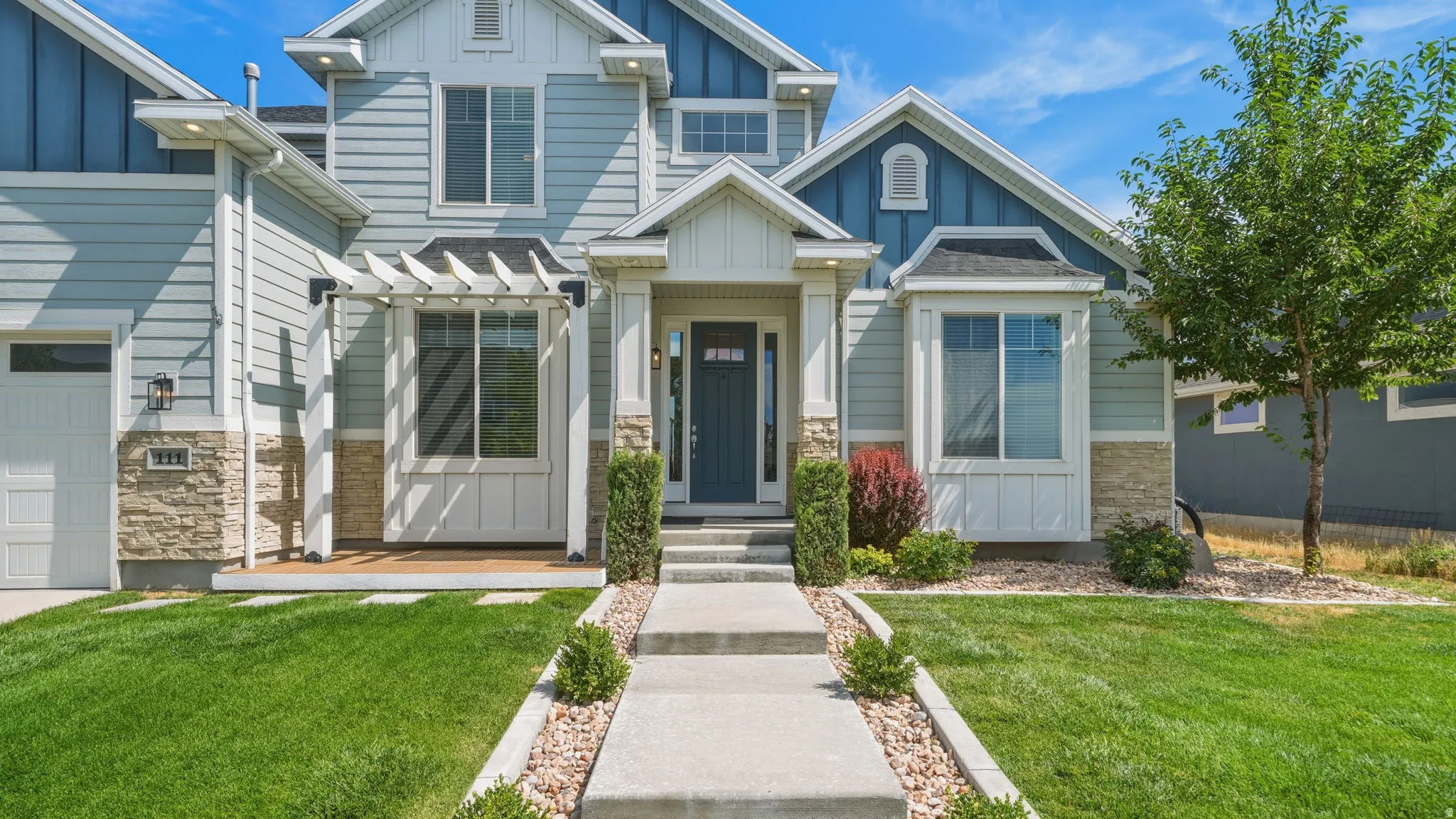Contemporary exterior featuring light blue siding, stone veneer accents, an inviting covered entry, a prominent bay window, and a neatly manicured lawn with a concrete pathway