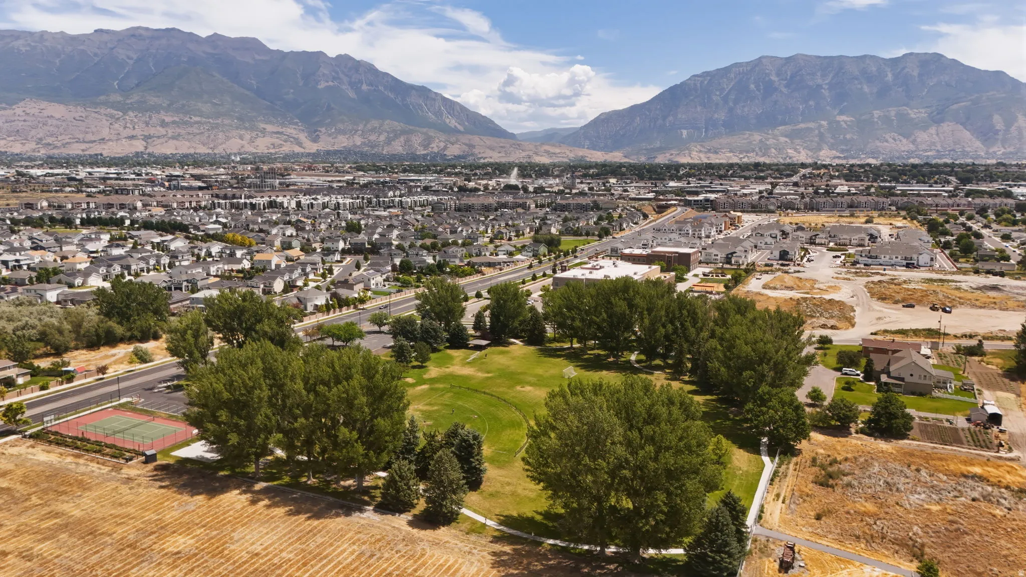 Expansive aerial perspective showcasing a neighborhood with diverse housing, mature trees, a manicured green space, and a red-surfaced sports court