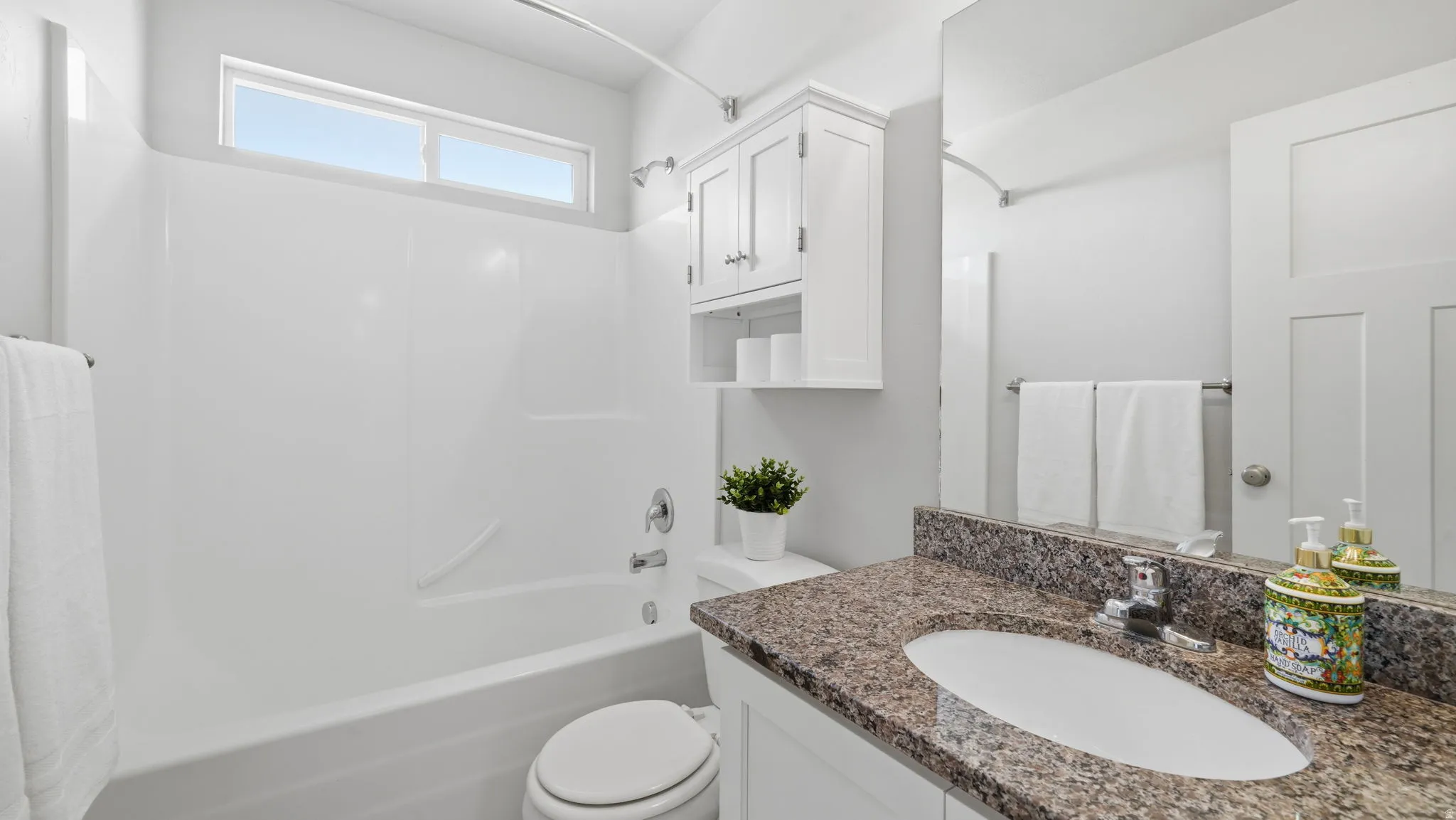 Bathroom featuring a white bathtub with an overhead transom window, a white toilet, and a vanity with a granite-style countertop, undermount sink, and white cabinetry
